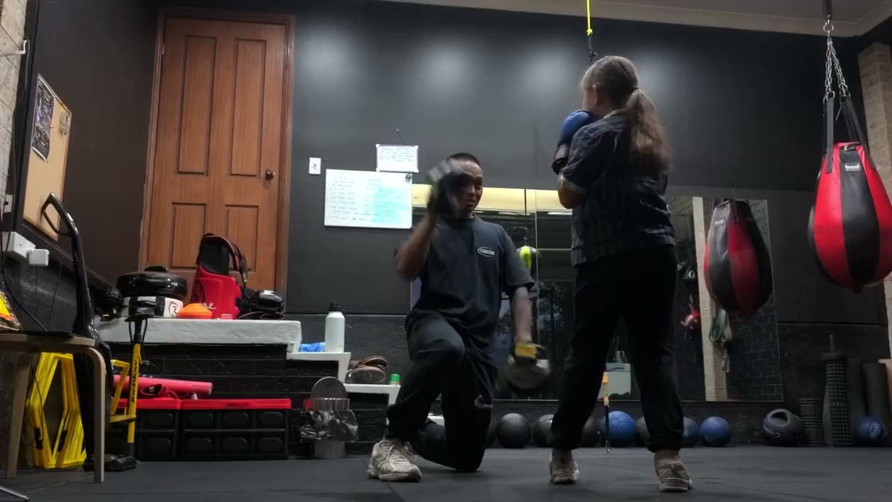 A coach kneeling with focus pads, coaching a young boxer through a combination in the Blitz gym.